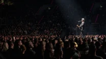 Tłum ludzi na widowni skupiony na scenie, gdzie samotny artysta w kapeluszu występuje z gitarą przy mikrofonie, a reflektor oświetla wykonawcę na tle ciemnej sali.