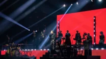 Grupa muzyków i wokalista na scenie koncertowej, jasne światła sceniczne oraz duży czerwony ekran w tle, widownia w półmroku