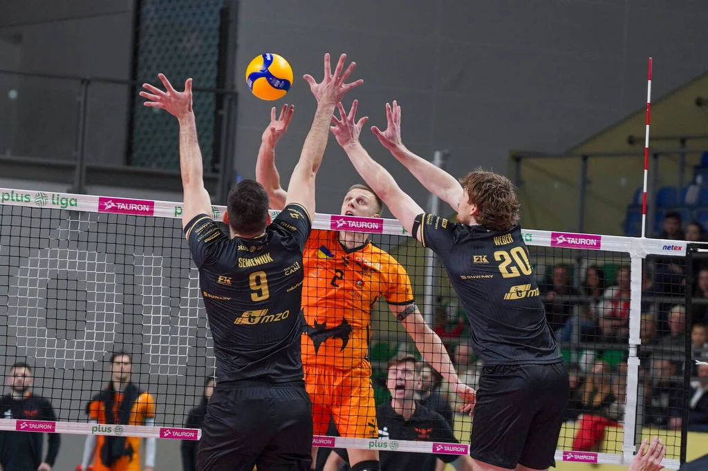 Blok PGE Projektu Warszawa Dwóch siatkarzy drużyny w czarnych strojach próbuje zablokować atak zawodnika ubranej w pomarańczowy strój, piłka znajduje się tuż nad siatką, wokół widoczni są inni zawodnicy oraz kibice na trybunach.