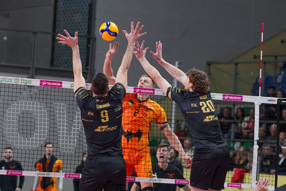 Barkom Każany Lwów wygrał w tym sezonie m.in. z PGE Projektem Warszawa Dwóch siatkarzy drużyny w czarnych strojach próbuje zablokować atak zawodnika ubranej w pomarańczowy strój, piłka znajduje się tuż nad siatką, wokół widoczni są inni zawodnicy oraz kibice na trybunach.