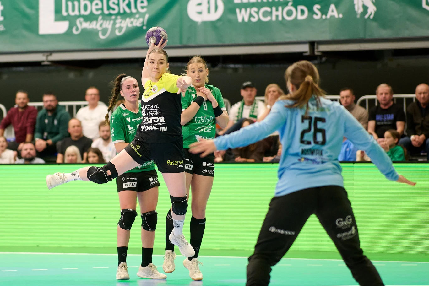 Zawodniczka drużyny w czarnej koszulce wykonuje rzut do bramki w trakcie meczu piłki ręcznej, podczas gdy dwie zawodniczki w zielonych koszulkach próbują ją zatrzymać, a bramkarka w jasnoniebieskiej bluzie przygotowuje się do obrony