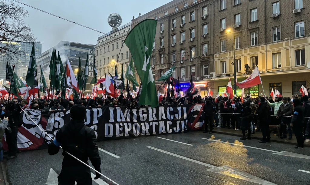 Tłum ludzi maszeruje ulicą miasta trzymając transparenty oraz flagi, z przodu widoczny duży napis Stop imigracji deportacje, w tle wysokie budynki oraz liczne zielone i biało-czerwone flagi.