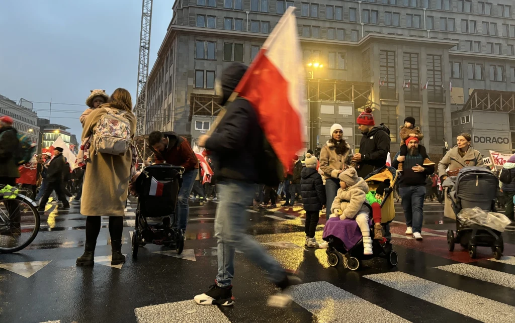 Tłum ludzi na dużym skrzyżowaniu w centrum miasta, wiele osób trzyma polskie flagi, wśród uczestników są rodzice z dziećmi w wózkach, panuje podniosła atmosfera patriotyczna, otoczenie budynków i lekka mgła.