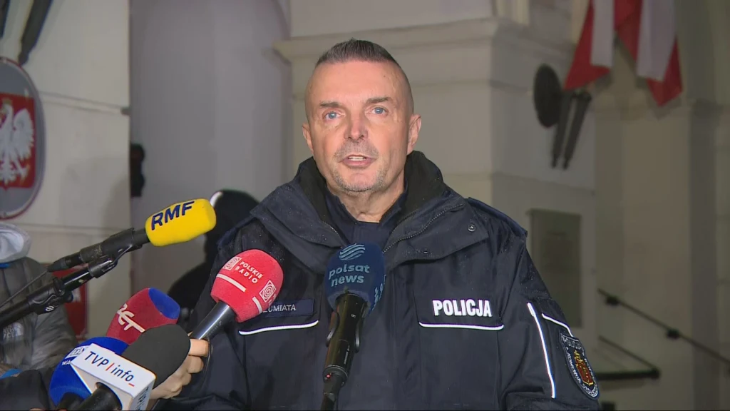 Rzecznik stołecznej policji podsumował Marsz Niepodległości Mężczyzna w policyjnym mundurze udzielający wypowiedzi przy mikrofonach różnych stacji telewizyjnych, na tle budynku z biało-czerwonymi flagami.