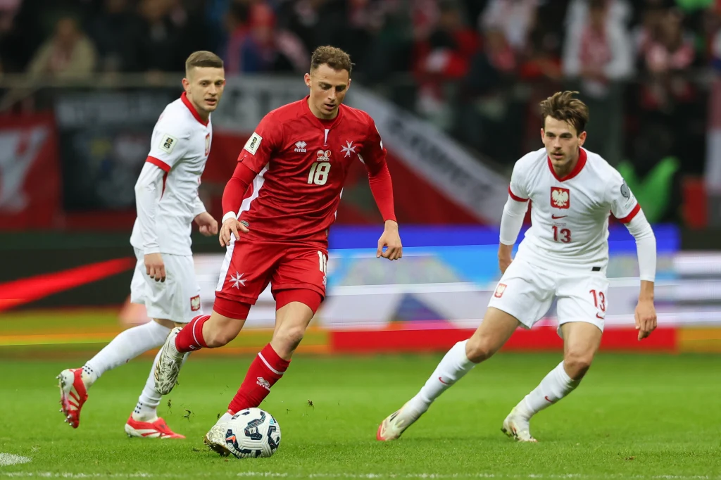 Piłkarze w trakcie meczu, zawodnik w czerwonym stroju prowadzi piłkę, a dwaj piłkarze w białych strojach obserwują sytuację i przygotowują się do interwencji.