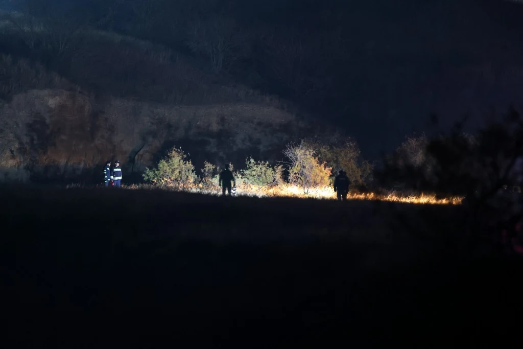 Kilka sylwetek ludzi poruszających się po ciemnej przestrzeni, oświetlonej silnym światłem, na tle zarośli i wzgórz nocą.