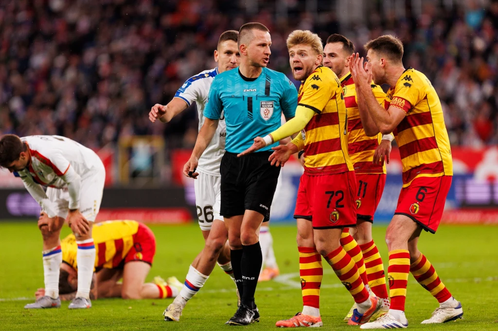 Mecz Górnik - Jagiellonia. To właśnie wtedy sędzia Frankowski podpadł kibicom z Białegostoku Sędzia piłkarski otoczony przez kilku piłkarzy w żółto-czerwonych strojach, którzy wyrażają swoje niezadowolenie lub protestują przeciwko jego decyzji. W tle inni zawodnicy, jeden z nich klęczy na murawie.