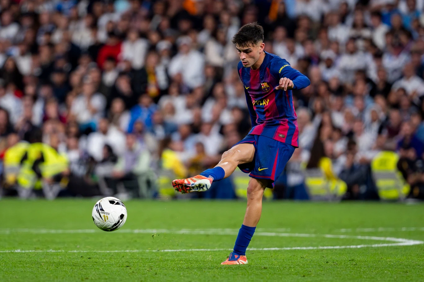 Piłkarz w niebiesko-bordowym stroju FC Barcelony wykonuje dynamiczny strzał piłki na boisku podczas meczu piłkarskiego, w tle widoczny tłum kibiców na trybunach.