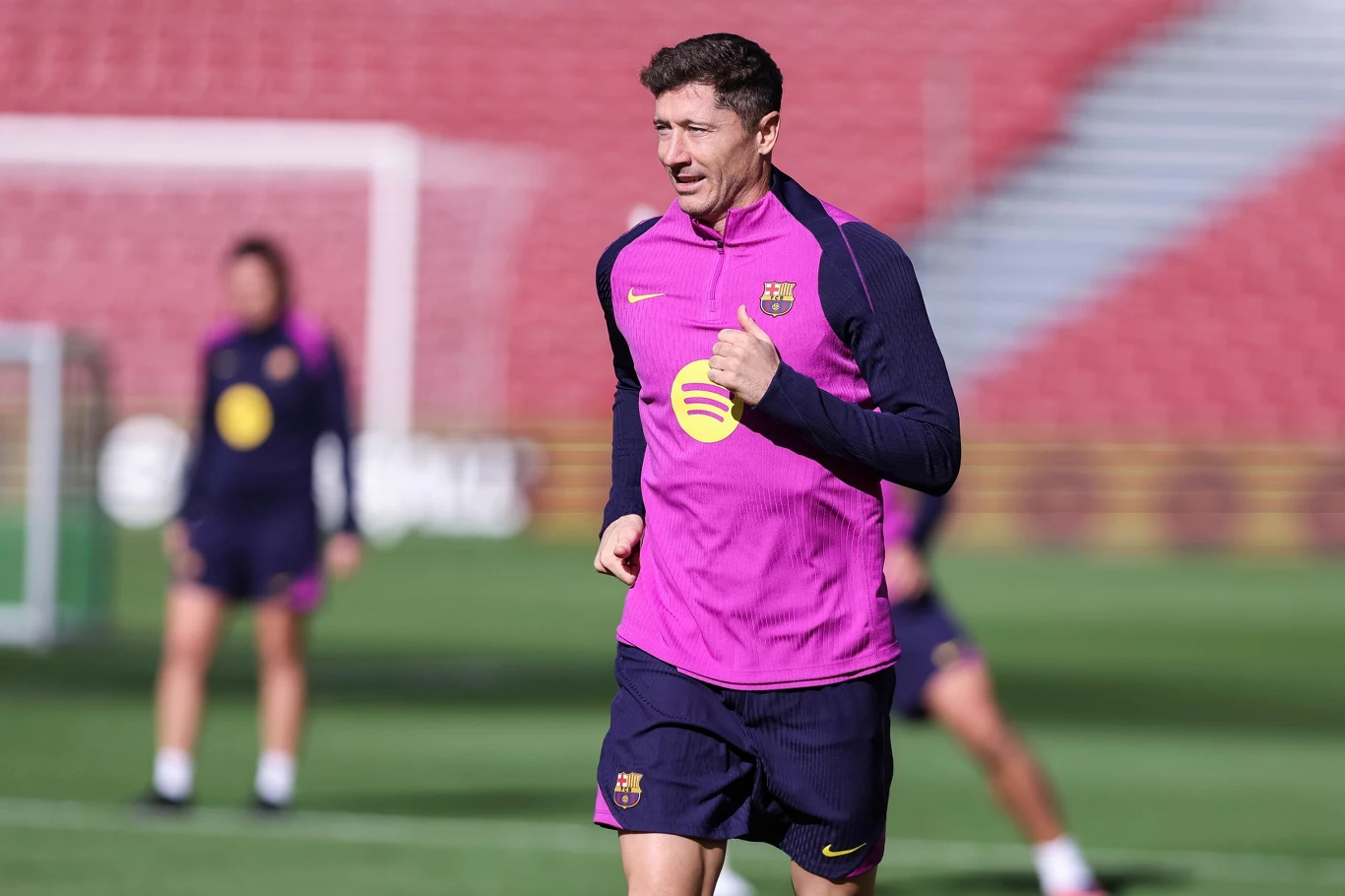 Piłkarz w stroju treningowym FC Barcelony biegnie po murawie stadionu podczas treningu, w tle widoczni są inni zawodnicy i rozmyte trybuny.