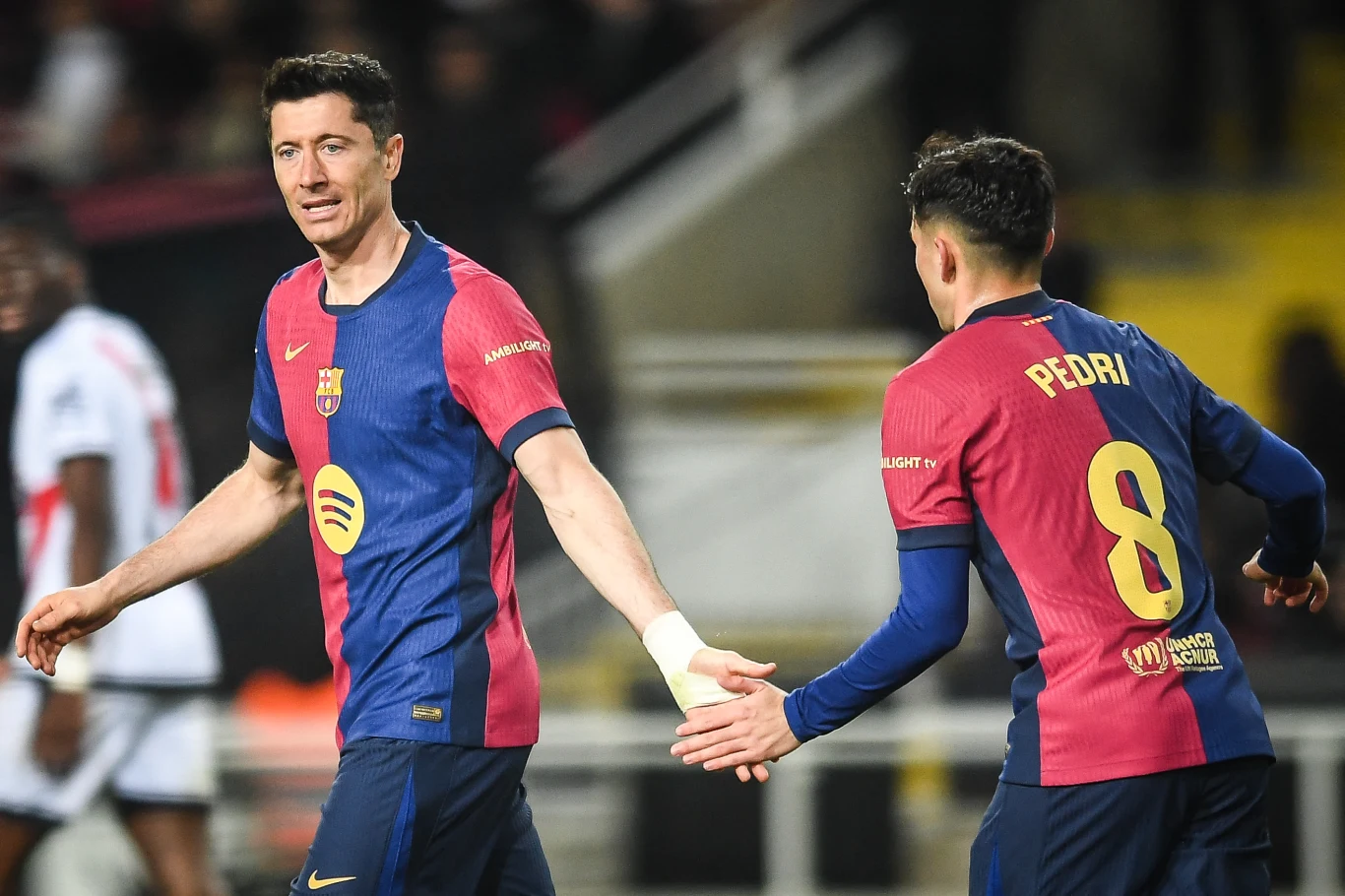 Dwóch piłkarzy drużyny FC Barcelona w granatowo-bordowych koszulkach podczas meczu, podają sobie ręce. Najważniejszymi elementami są wyrażenia twarzy oraz gesty zawodników, a także widoczne nazwiska i numery na plecach jednego z nich.