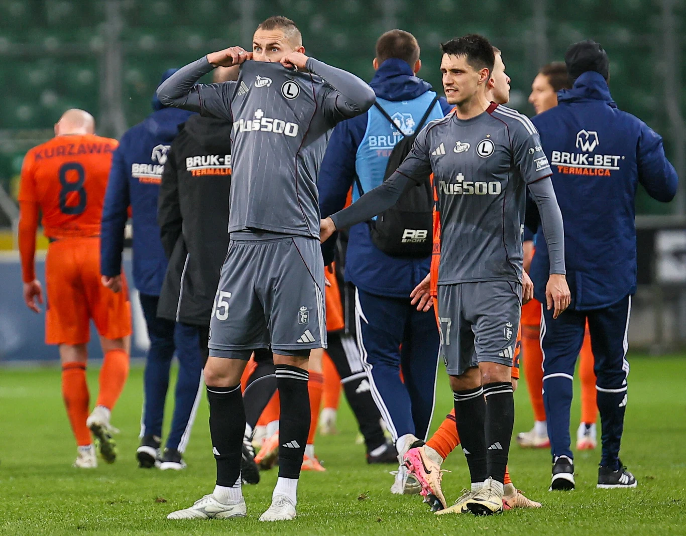 Legia Warszawa zajmuje aktualnie dopiero 11. miejsce w tabeli Ekstraklasy Piłkarze dwóch drużyn na boisku po zakończonym meczu, zawodnicy w szarych strojach wyglądają na rozczarowanych, jeden z nich zakrywa twarz koszulką. W tle piłkarze w pomarańczowych strojach oraz sztab szkoleniowy.