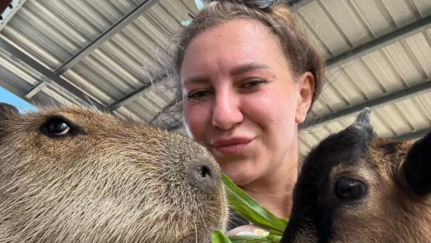 Kobieta robi selfie, karmiąc dużą kapibarę i kozę świeżą zieloną rośliną pod zadaszeniem.