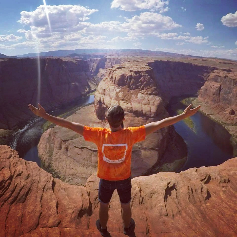 Mateusz Kowalewicz Osoba ubrana w pomarańczową koszulkę stoi z rozłożonymi ramionami na krawędzi skalnego urwiska nad zakolem rzeki, w tle rozciąga się szeroki kanion pod błękitnym niebem z lekkimi chmurami.
