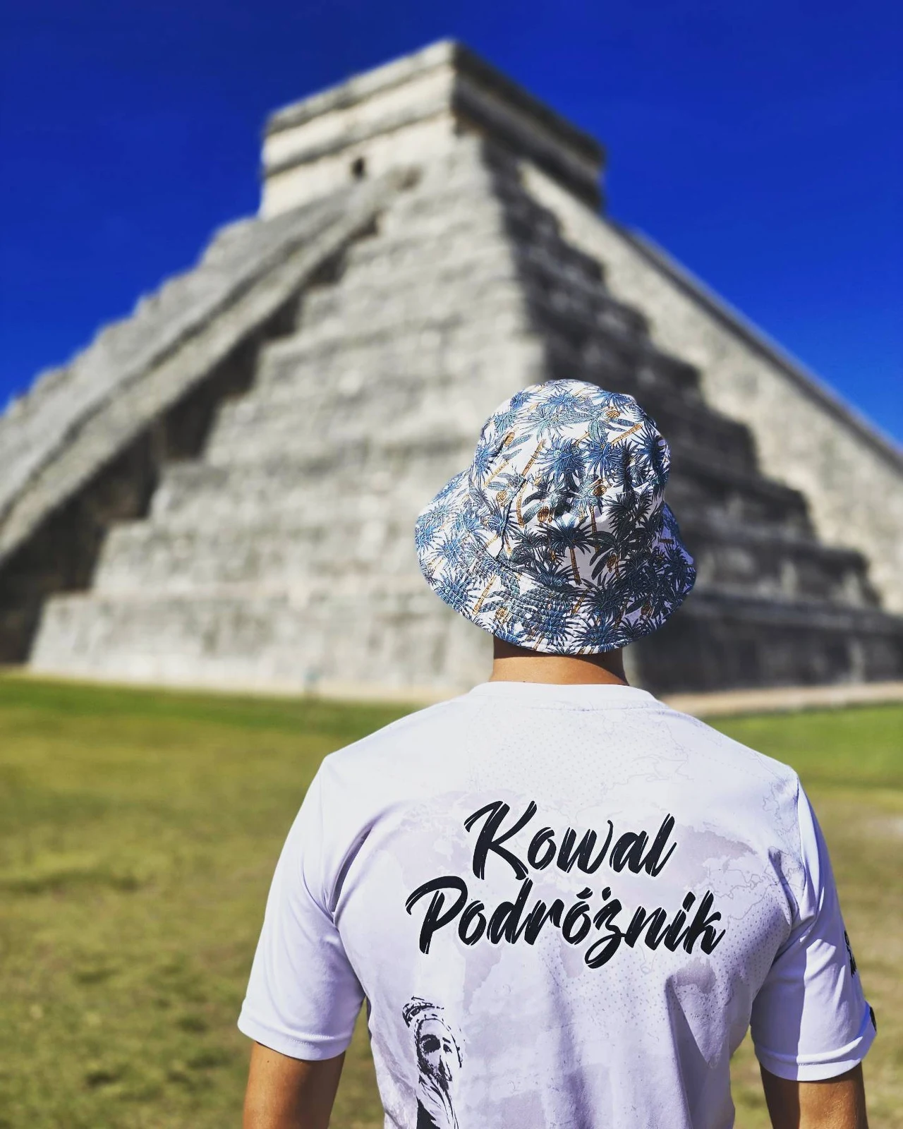Mateusz Kowalewicz Mężczyzna w kapeluszu i białej koszulce z napisem 'Kowal Podróżnik' stoi tyłem do monumentalnej piramidy Majów, która wyróżnia się na tle nieba.