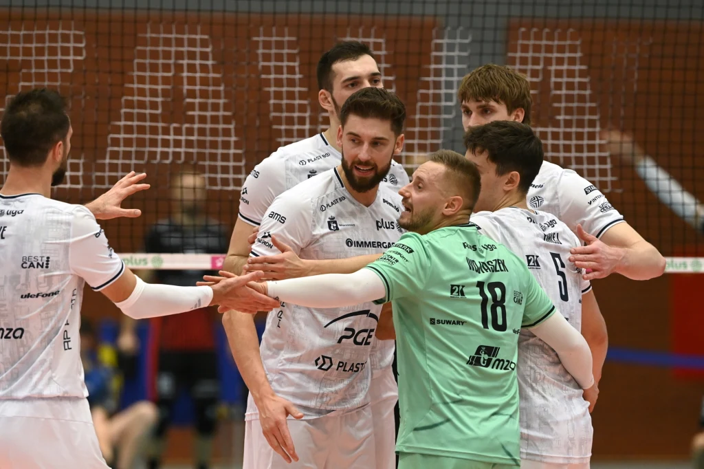 Siatkarze PGE Projektu Warszawa Grupa siatkarzy ubranych w białe stroje oraz jeden zawodnik w zielonej koszulce cieszy się ze zdobytego punktu podczas meczu siatkarskiego, wszyscy znajdują się tuż przy siatce na hali sportowej.