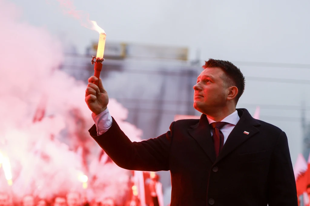 11 listopada. Sławomir Mentzen złamał zakaz dotyczący rac na Marszu Niepodległości Mężczyzna w garniturze trzymający zapaloną racę w tłumie ludzi otoczonych czerwonym światłem od rac i dymem, tło rozjaśnione przez intensywne światło pochodni, wyraz twarzy poważny i skupiony.