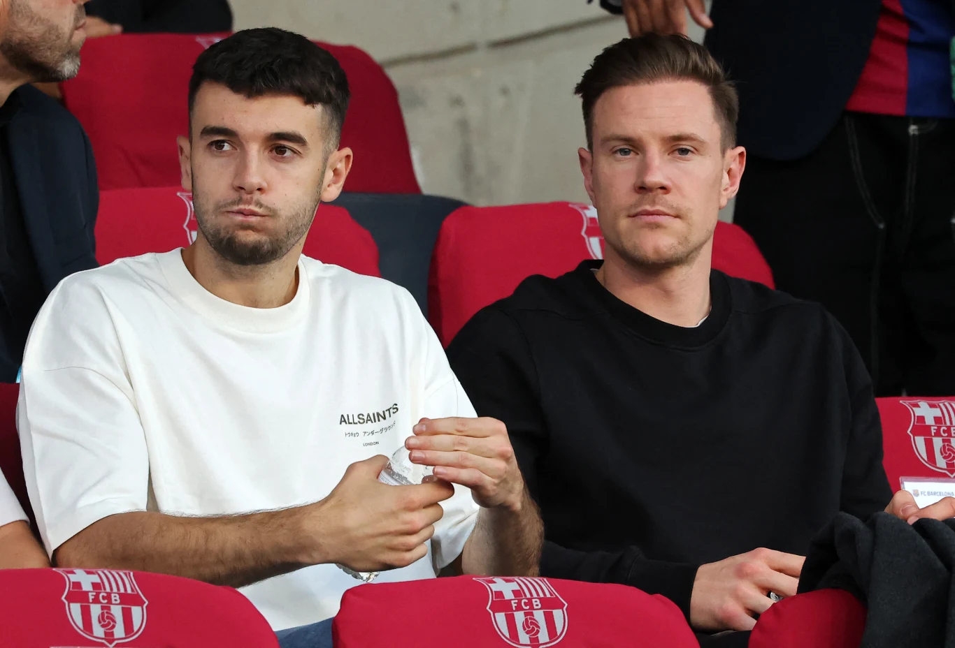 Joan Garcia, Marc-Andre ter Stegen Dwóch mężczyzn siedzących obok siebie na czerwonych fotelach stadionowych, jeden w białej koszulce, drugi w czarnej. Obaj patrzą przed siebie, a jeden z nich trzyma coś w rękach.