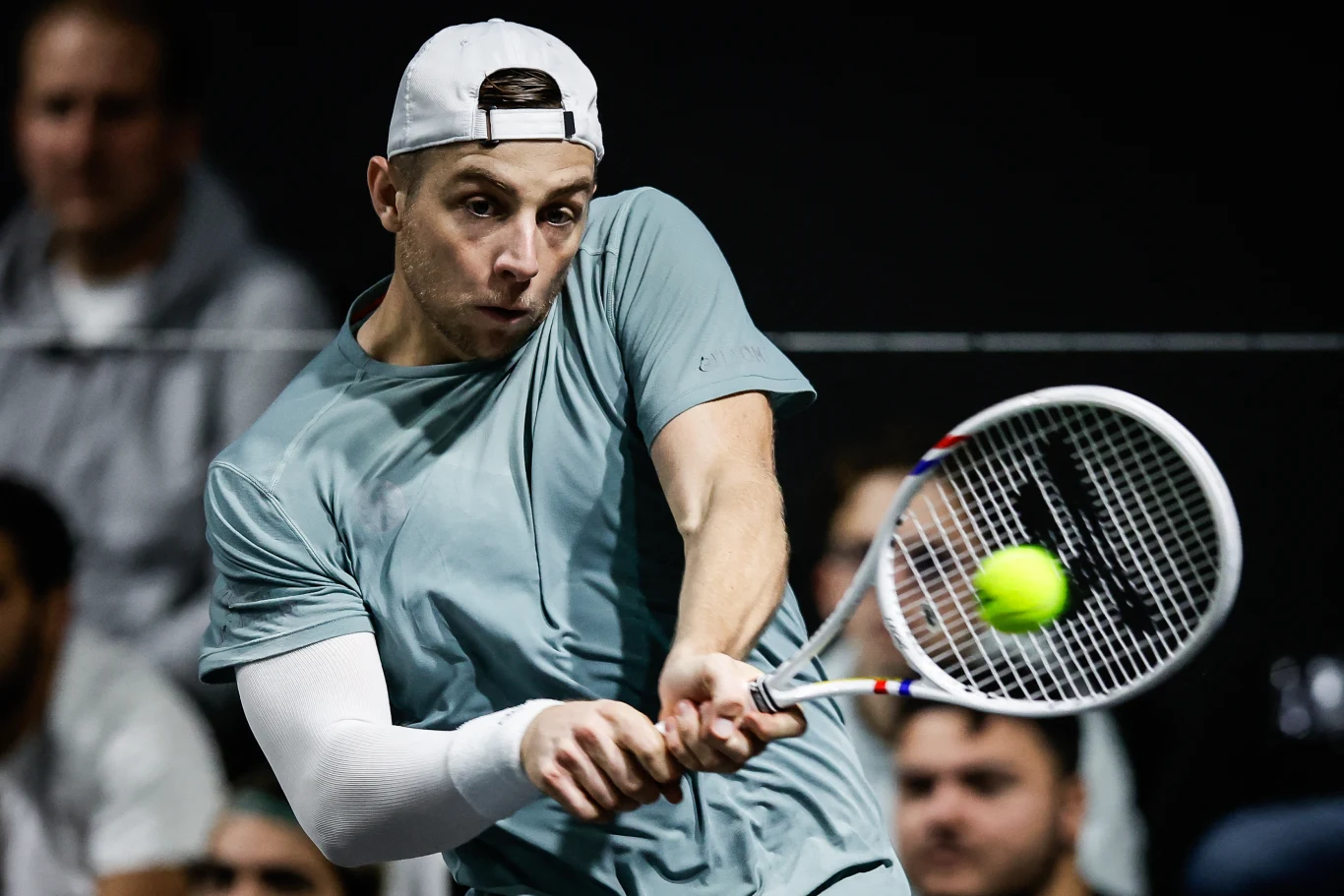 Tenisista w czapce uderza piłkę rakietą podczas dynamicznego meczu, skupiony na grze, w tle widoczna publiczność.