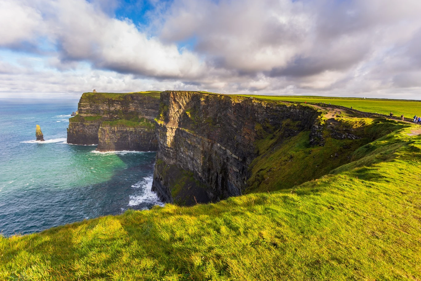 Zielone klify wznoszą się wysoko nad oceanem, u ich podnóża fale rozbijają się o skały. Po prawej stronie widoczna ścieżka prowadzi wzdłuż krawędzi, w tle rozległe niebo z chmurami.