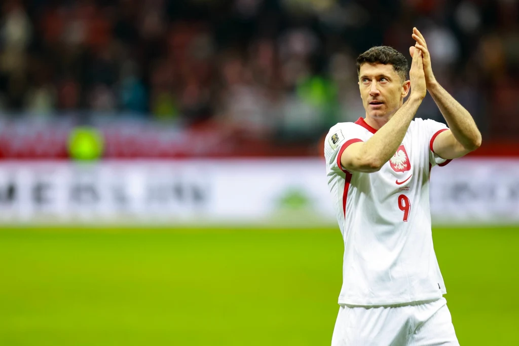 Piłkarz w białym stroju z numerem 9 na koszulce bije brawa na stadionie, na tle rozmytej publiczności i jasnych świateł.