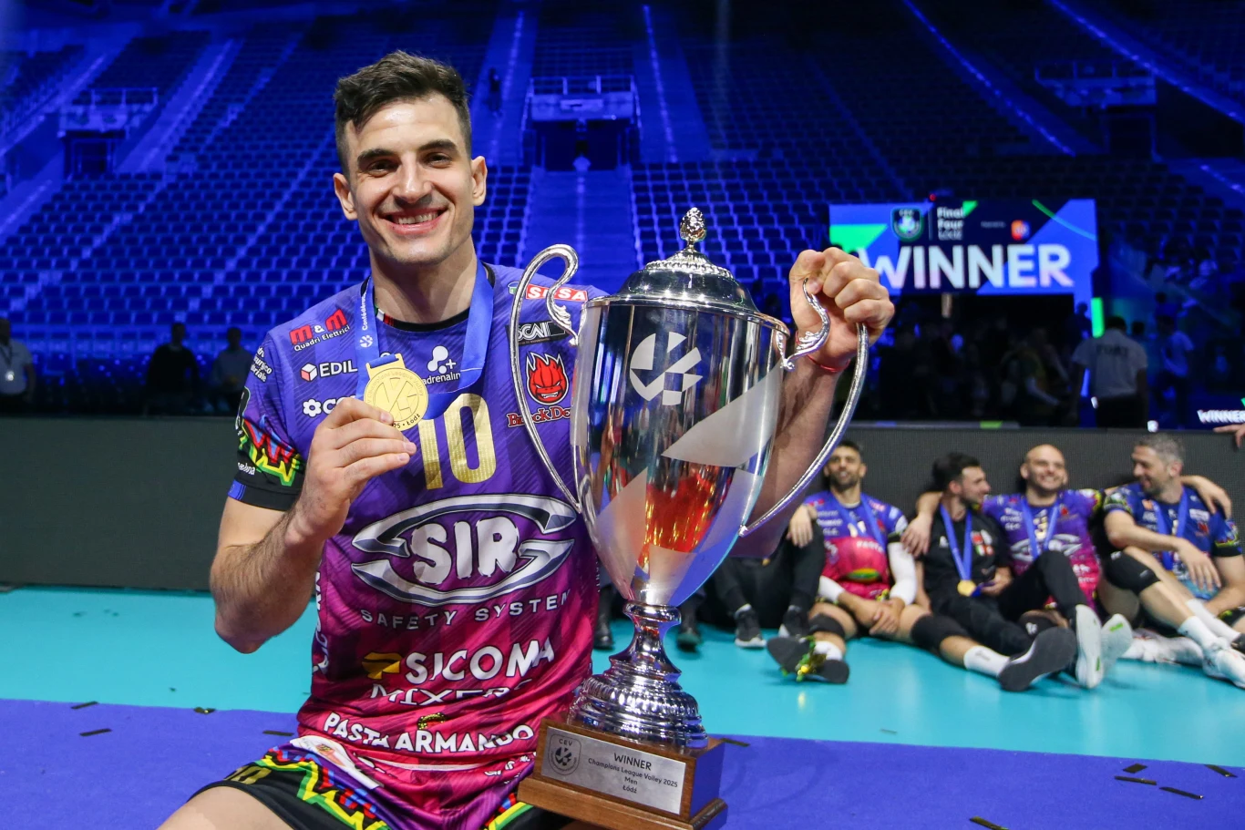 Zawodnik drużyny siatkarskiej w kolorowej koszulce trzyma duży puchar i złoty medal, szeroko się uśmiecha; w tle na niebieskim parkiecie siedzi grupa współzawodników i wyraźnie widoczny jest napis 'WINNER'.