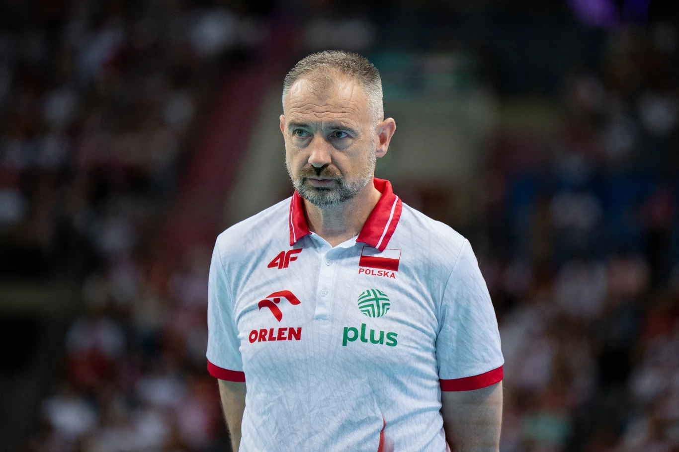 Mężczyzna w sportowej koszulce z logo sponsorów i napisem Polska stoi na tle rozmytej publiczności podczas wydarzenia sportowego.