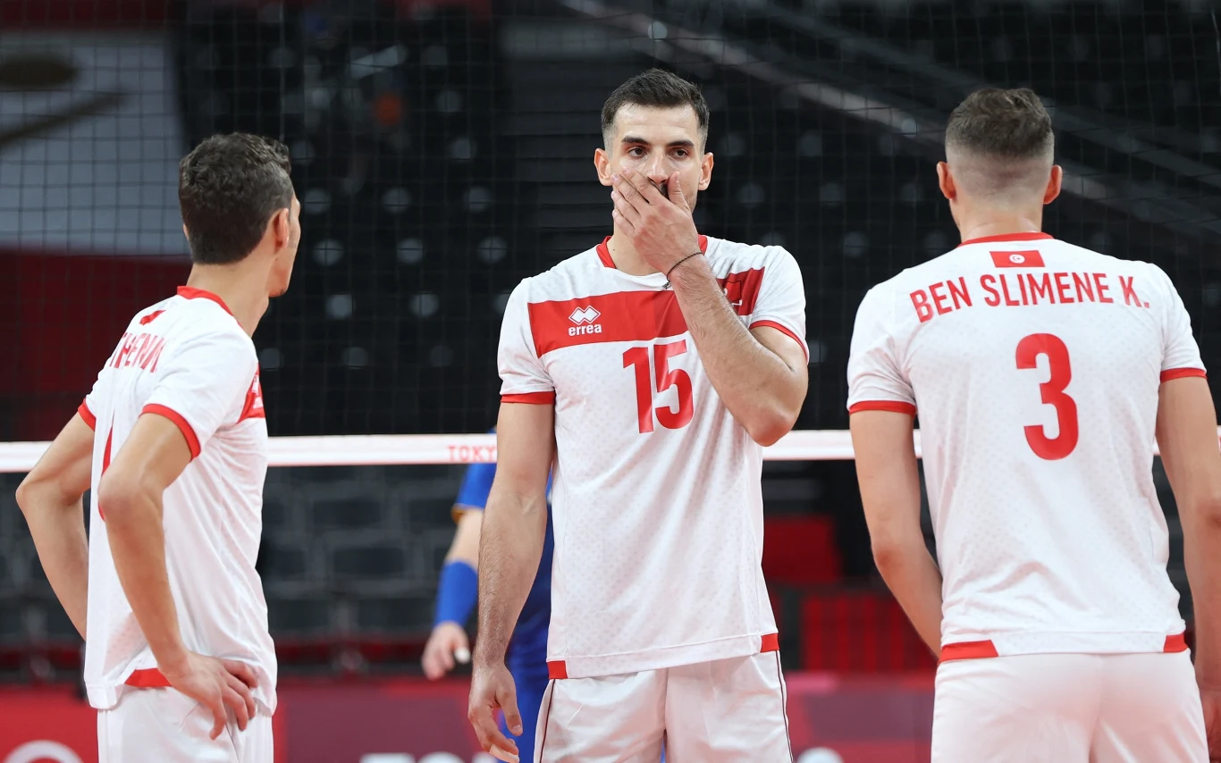 Wassim Ben Tara w barwach reprezentacji Tunezji Trzech siatkarzy w białych strojach z czerwonymi akcentami stoi na boisku, dwóch z nich widocznych z tyłu, jeden z przodu zasłania dłonią usta. W tle siatka i puste trybuny.