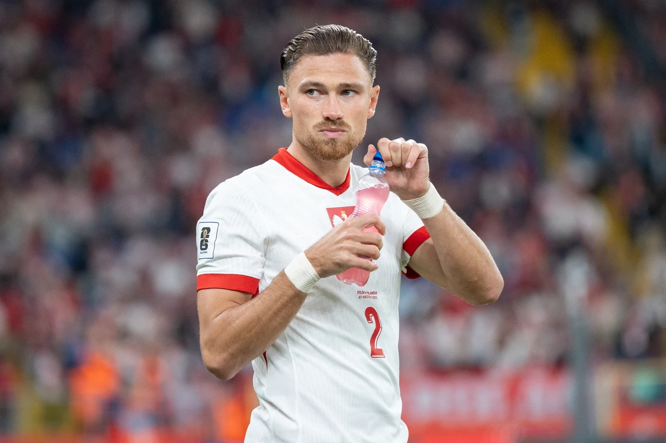 Piłkarz w białej koszulce z numerem 2 i godłem Polski trzyma butelkę z napojem, stojąc na boisku podczas meczu, w tle zamazana publiczność i stadion.