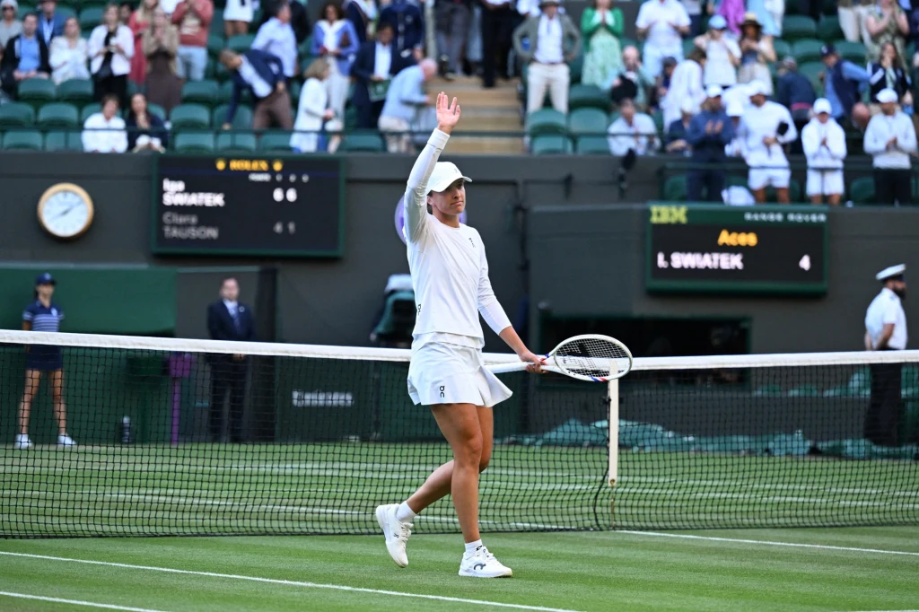 Iga Świątek po jednym z setów wygranych 6:1. Z Clarą Tauson w Wimbledonie Tenisistka w białym stroju stoi na korcie, trzyma rakietę w jednej ręce i unosi drugą, machając do publiczności. W tle trybuny z widzami, tablice z wynikami oraz sędziowie i obsługa.