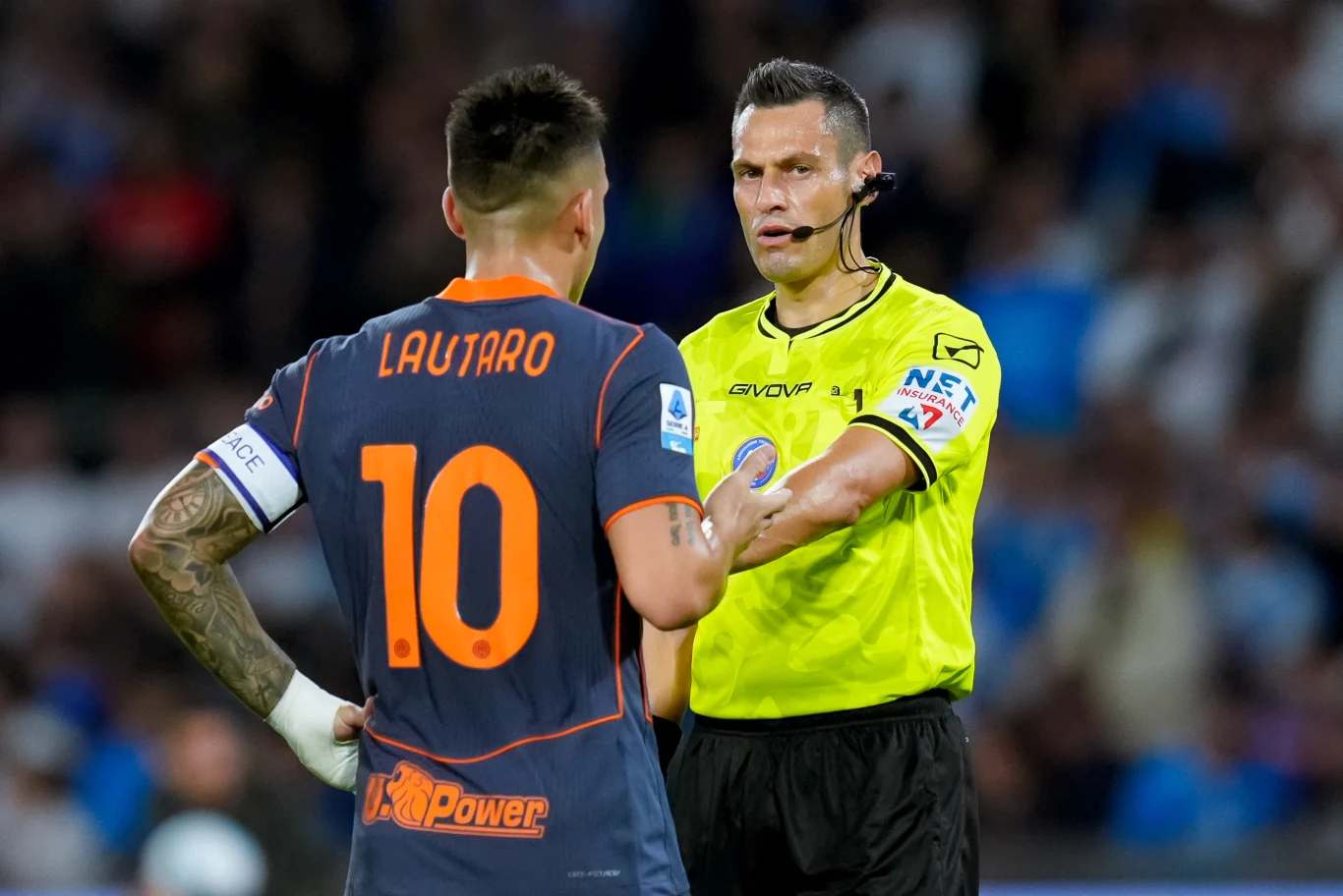 Sędzia piłkarski w żółtej koszulce rozmawia z piłkarzem o nazwisku Lautaro, który nosi ciemny strój z numerem 10 na plecach. Scena rozgrywa się na tle stadionu pełnego widzów.