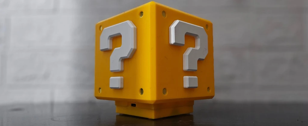 Najlepsze gry na Nintendo Switch 2 Żółty sześcian z trzema dużymi białymi znakami zapytania na ściankach, ustawiony na ciemnej powierzchni na tle rozmytej jasnoszarej ściany.