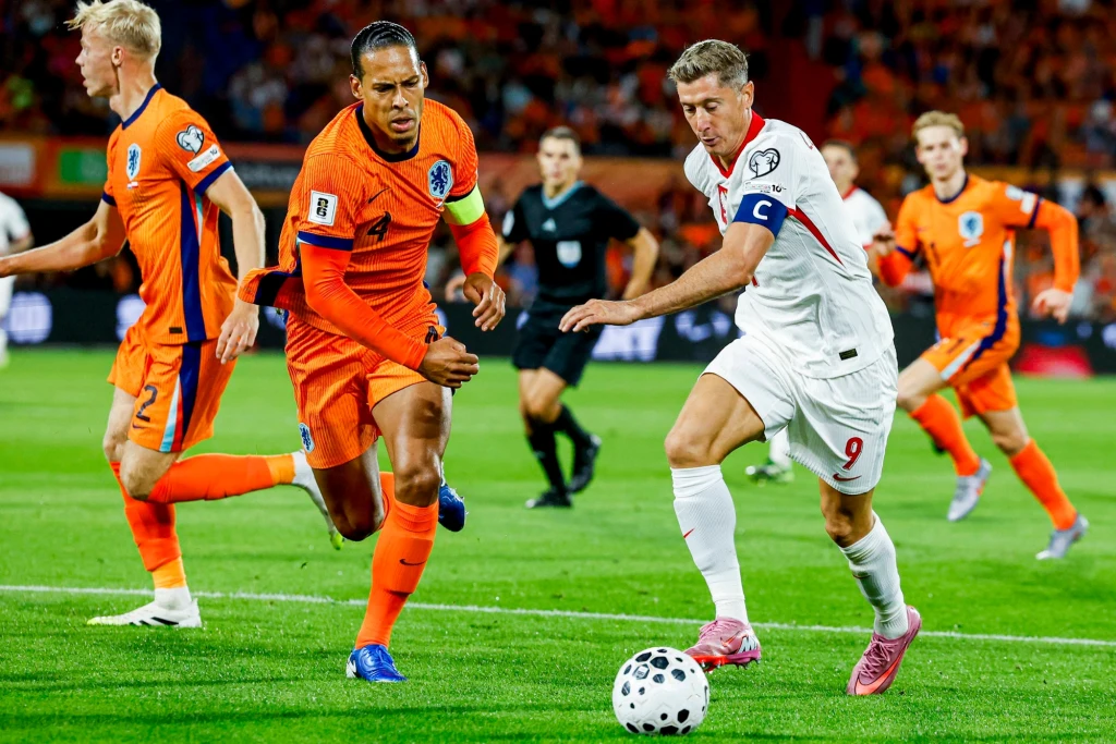 Robert Lewandowski w meczu Holandia - Polska 4.09.2025 Dwóch piłkarzy rywalizuje o piłkę podczas meczu reprezentacji Holandii z drużyną ubraną na biało. Obaj zawodnicy mają opaski kapitańskie na ramionach. W tle widoczni są sędziowie oraz inni piłkarze na boisku.