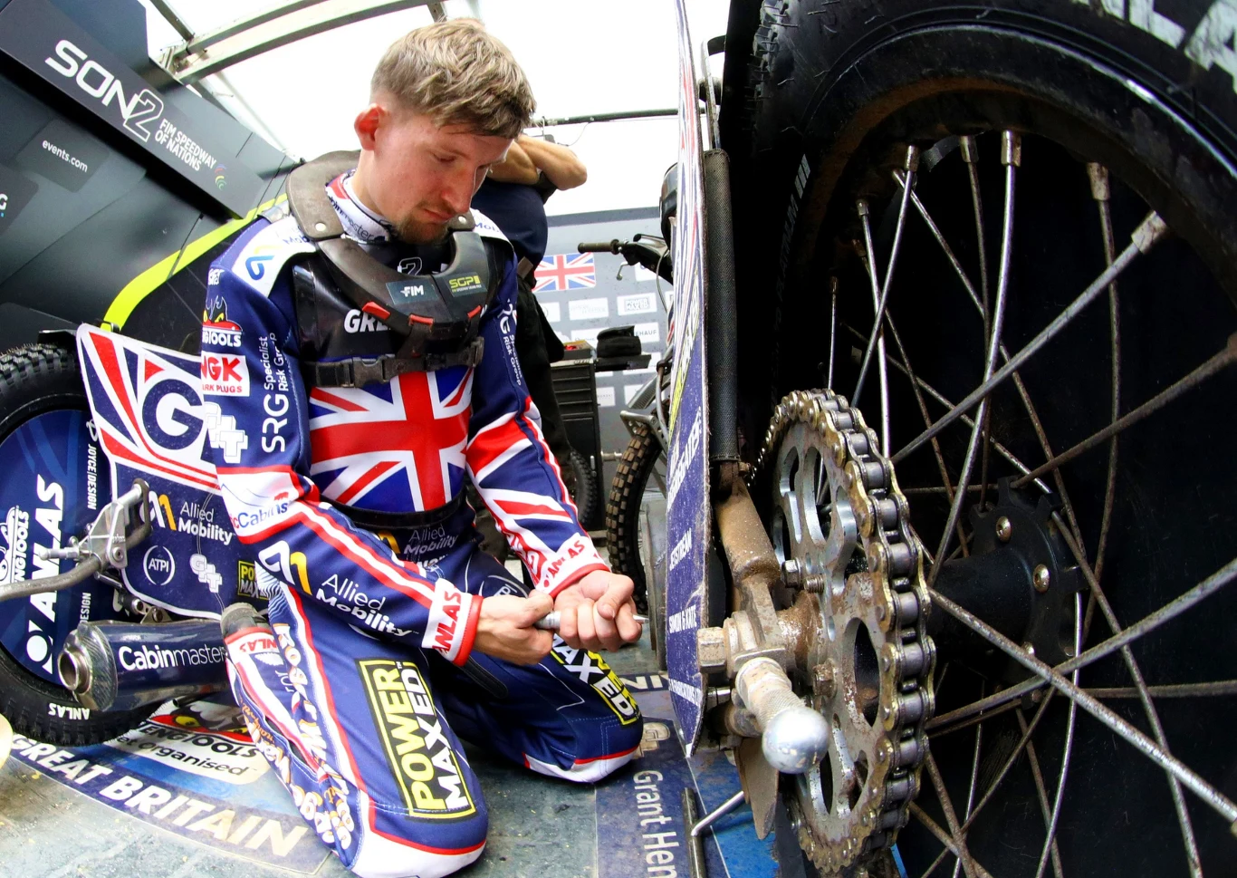 Zawodnik ubrany w strój z flagą Wielkiej Brytanii klęczy obok motocykla żużlowego, pracując przy tylnym kole oraz napędzie. Sylwetka sportowca skupiona jest na czynnościach serwisowych pojazdu.