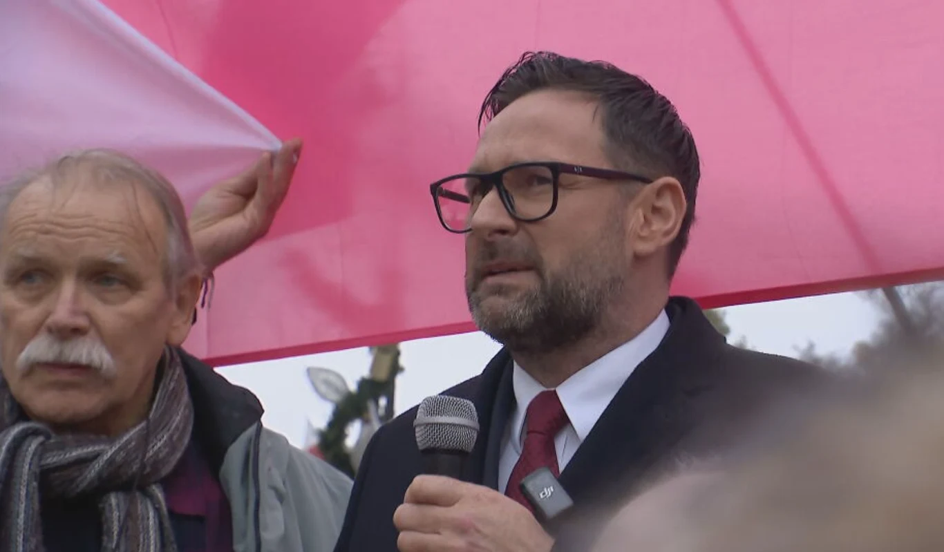 Daniel Obajtek zjawił się w prokuraturze. Przed wejściem wygłosił przemówienie Dwóch mężczyzn pod dużą biało-czerwoną flagą, jeden z nich trzyma mikrofon i wygłasza przemówienie, drugi przytrzymuje flagę. W tle widoczne delikatnie rozmyte elementy krajobrazu oraz fragmenty dekoracji.