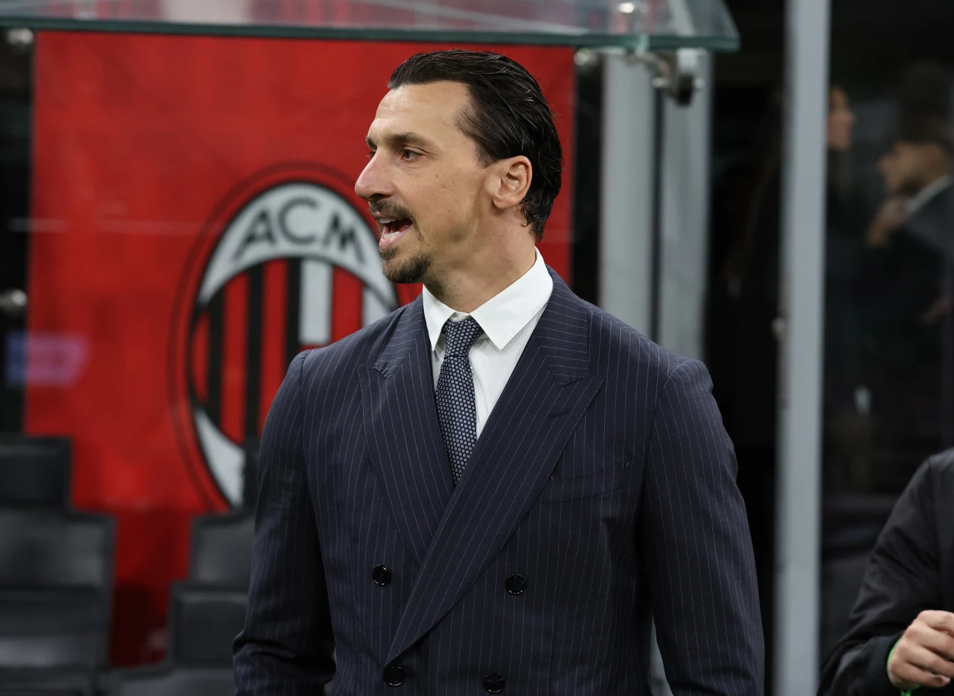 Mężczyzna w eleganckim, ciemnym garniturze z krawatem stoi przed czerwono-czarną flagą klubu piłkarskiego AC Milan w stadionowej loży.