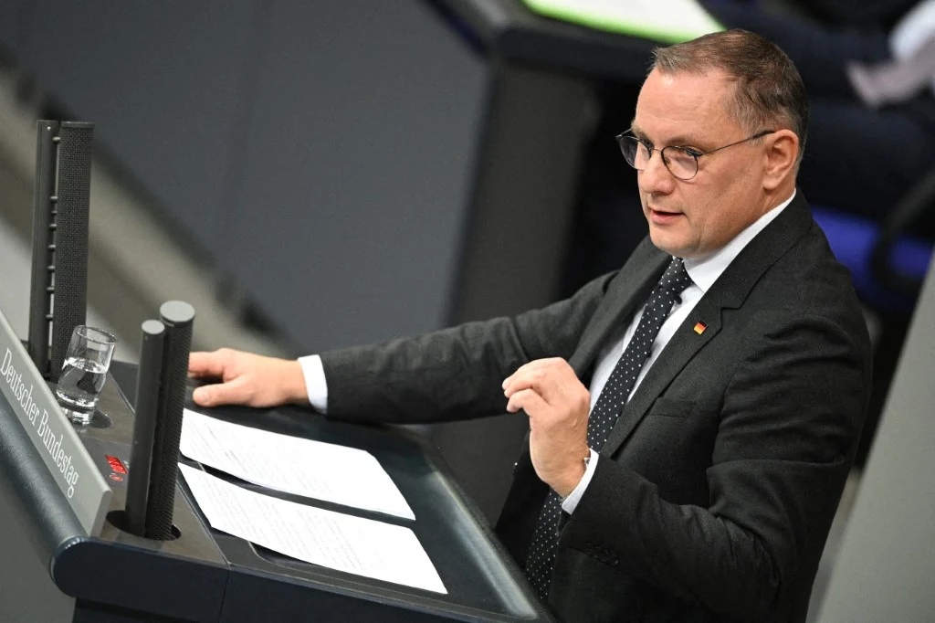 Współlider niemieckiej AfD Tino Chrupalla Mężczyzna w garniturze przemawia przy mównicy, przed nim leżą kartki z dokumentami, a obok znajduje się szklanka wody. Osoba wykonuje gest ręką, skupiona na przemówieniu.