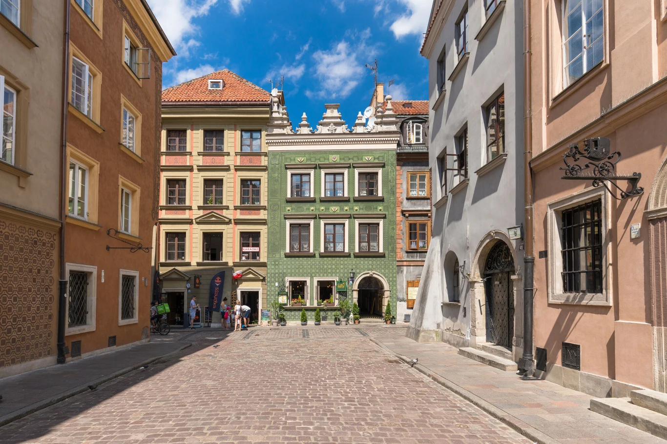 Wąska brukowana uliczka otoczona kolorowymi kamienicami, w centrum wyróżnia się zielona kamienica z bogato zdobioną fasadą, a po bokach widoczne są okna, drzwi i detale architektoniczne.