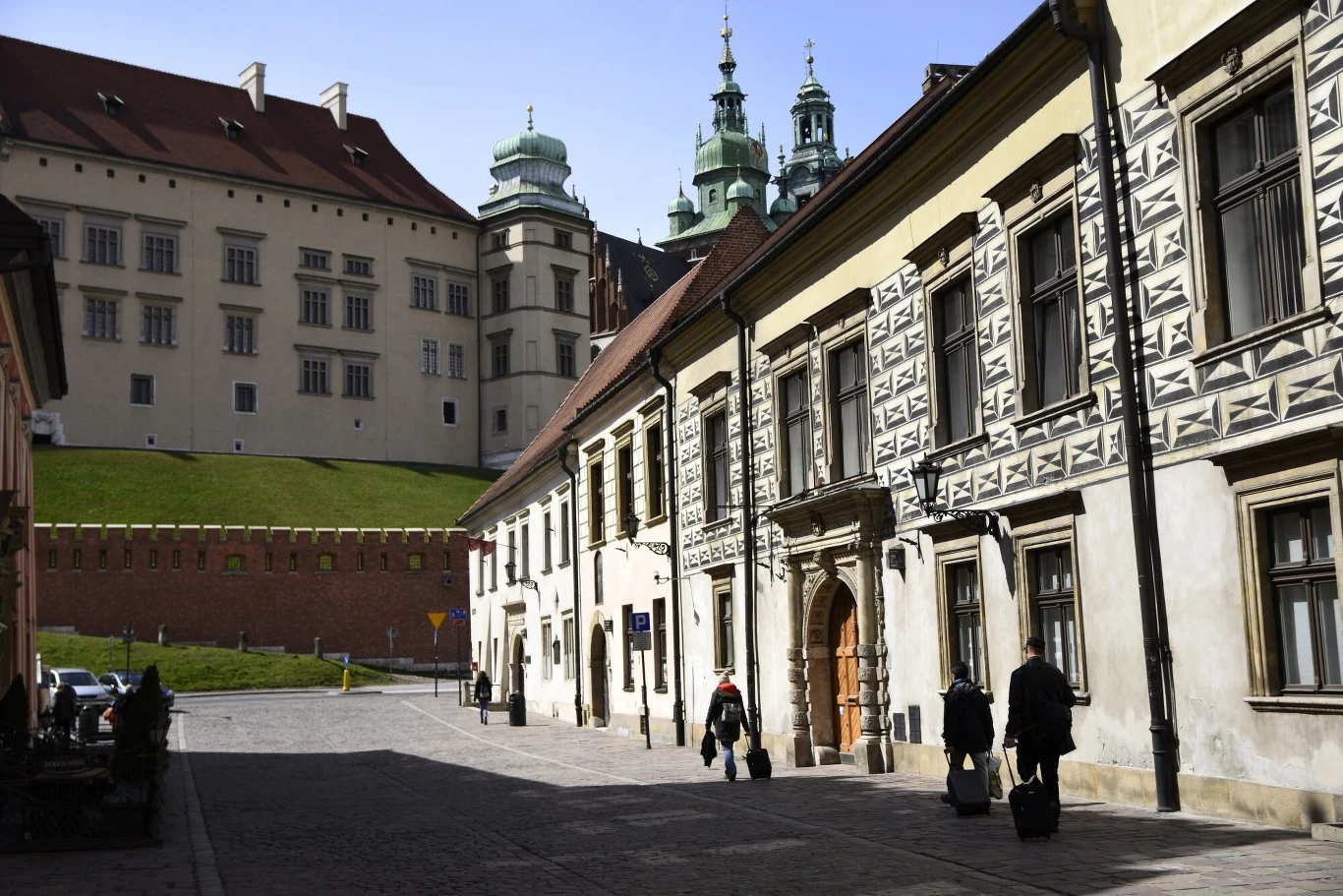 Historyczna ulica w Krakowie z charakterystycznymi renesansowymi kamienicami, widokiem na wzgórze Wawelskie i fragmentem murów zamkowych, po chodniku chodzą osoby z walizkami.