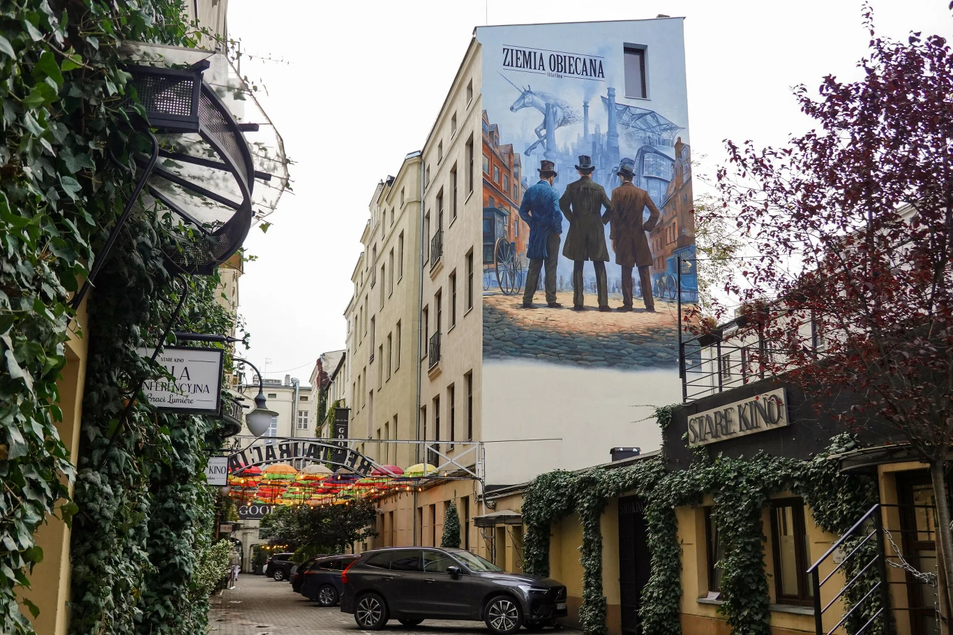 Wąska ulica w centrum miasta otoczona porośniętymi bluszczem budynkami, na bocznej ścianie jednego z nich duży mural z trzema mężczyznami ubranymi w stroje z przełomu XIX i XX wieku, z napisem 'Ziemia Obiecana'. Wzdłuż ulicy zaparkowany jest samochód, ...