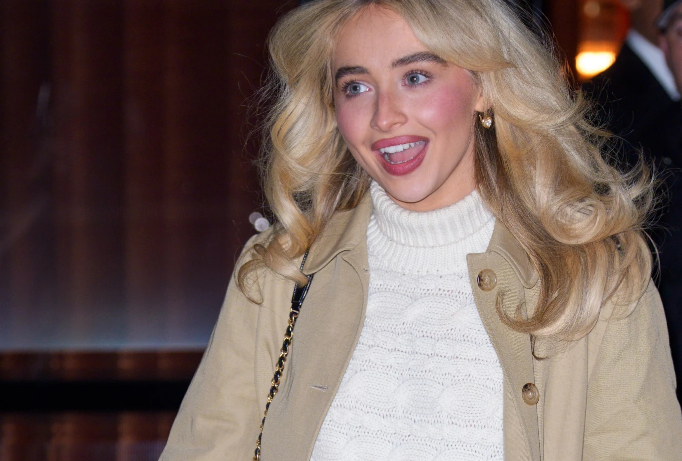 Sabrina Carpenter wystąpi w adaptacji "Alicji w Krainie Czarów Młoda kobieta z blond włosami ułożonymi w fale, ubrana w jasny sweter i beżowy płaszcz, uśmiechnięta i patrząca lekko w bok, złote kolczyki i torebka na ramieniu.