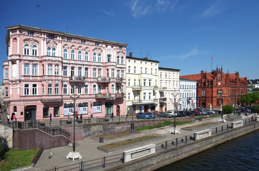 Rząd zabytkowych kamieniczek i czerwony gmach urzędu miejskiego przy nabrzeżu rzeki, jasne fasady budynków i czysta niebieska nieba, wzdłuż brzegu rozmieszczone ławki, latarnie oraz zaparkowane samochody.