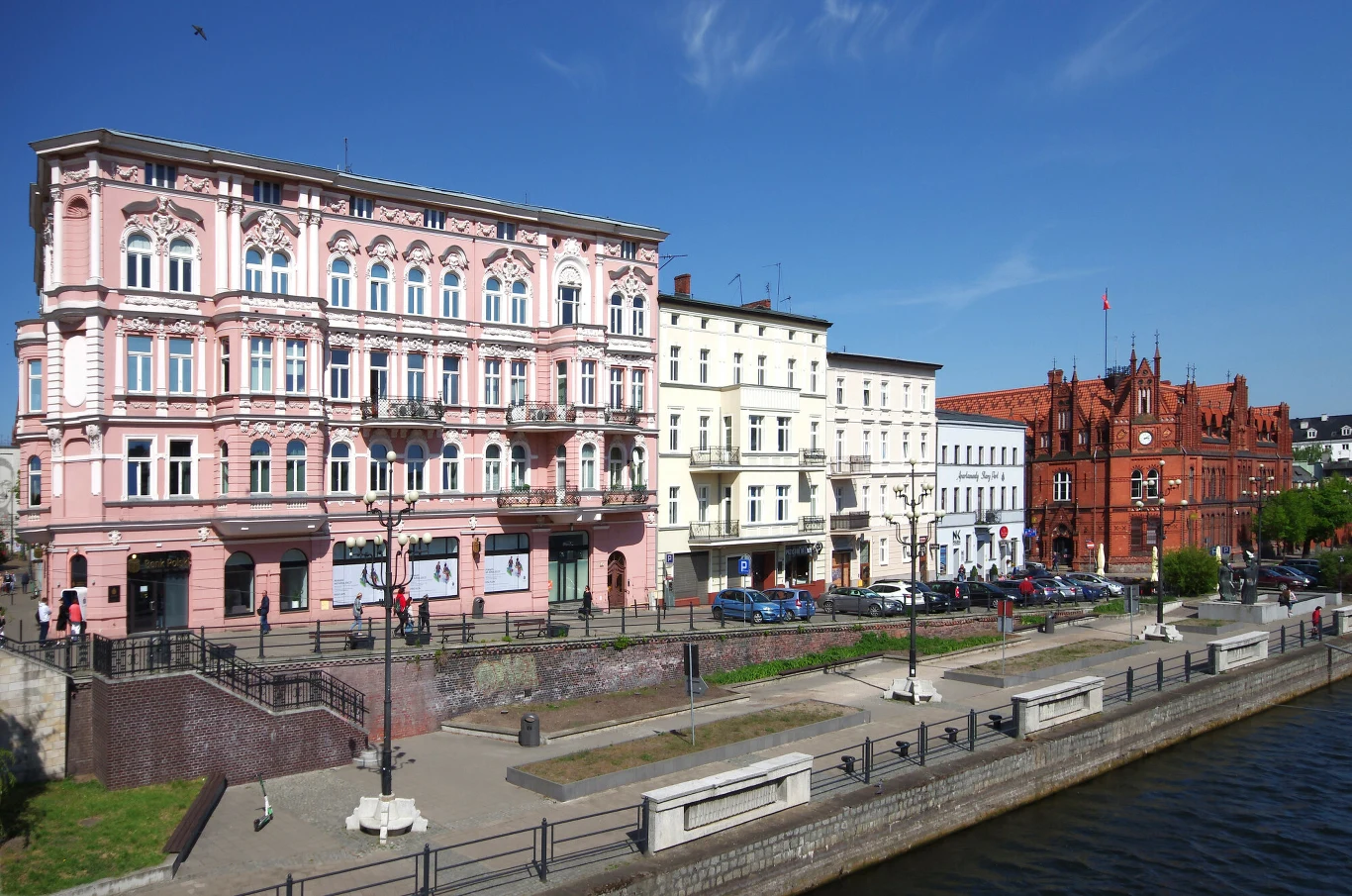 Rząd zabytkowych kamieniczek i czerwony gmach urzędu miejskiego przy nabrzeżu rzeki, jasne fasady budynków i czysta niebieska nieba, wzdłuż brzegu rozmieszczone ławki, latarnie oraz zaparkowane samochody.