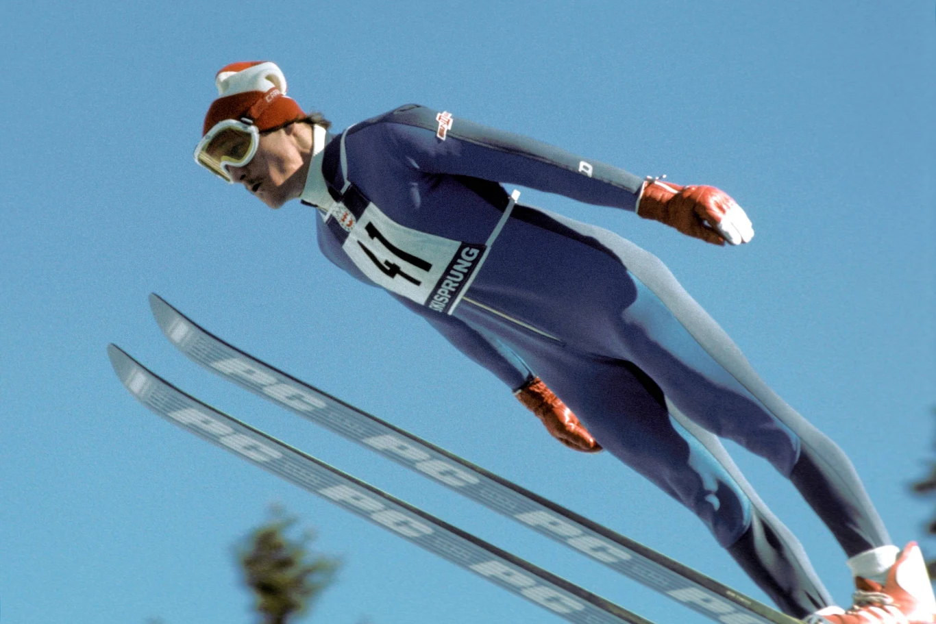 Sportowiec w granatowym kombinezonie oraz kasku ze śnieżnobiałą czapką unosi się w powietrzu podczas skoku narciarskiego, trzymając narty blisko siebie na tle jasnoniebieskiego nieba.