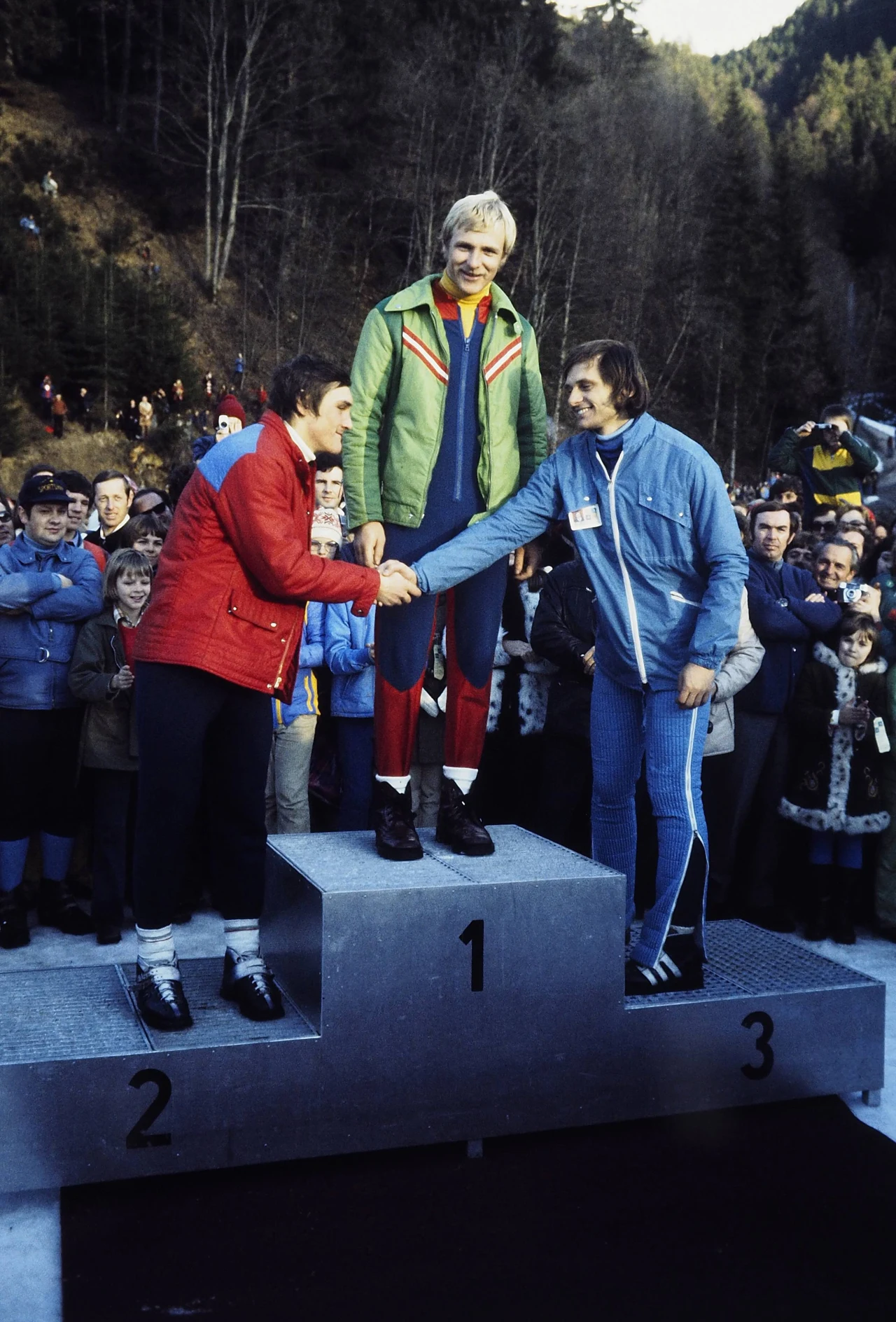 Trzech sportowców stoi na podium, zwycięzca na najwyższym stopniu otrzymuje gratulacje od pozostałych zawodników, wokół zgromadzony tłum obserwatorów na tle górskiego krajobrazu.