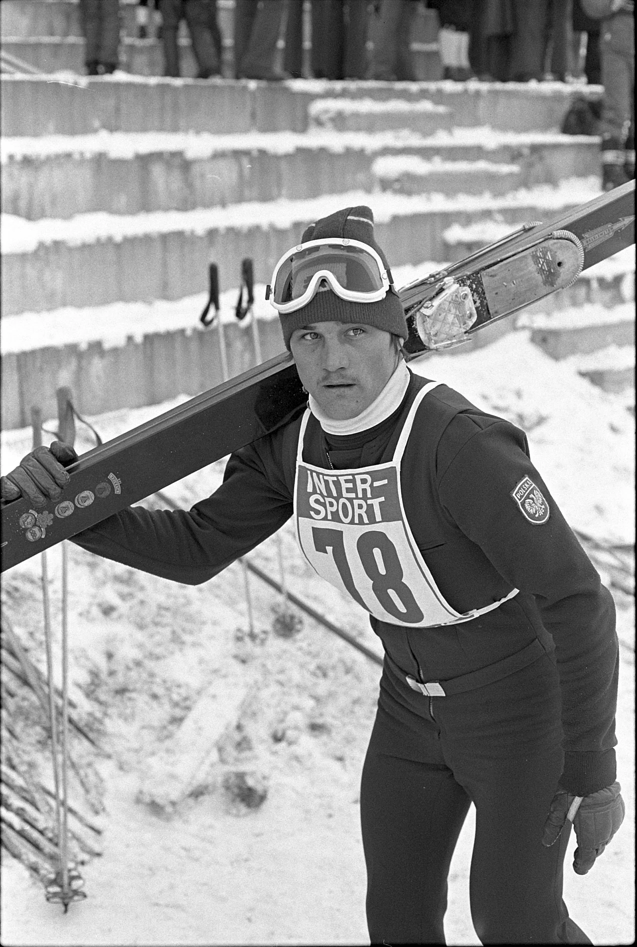 Narciarz ubrany w sportowy kombinezon z numerem startowym 78 niesie narty na ramieniu, w tle widoczne są zimowe schody i inni uczestnicy lub kibice wydarzenia.