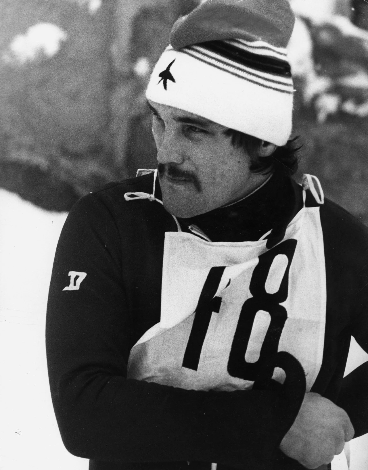 Narciarz w sportowym stroju zimowym z numerem startowym 18 oraz czapką z gwiazdą, stojący na tle ośnieżonego krajobrazu.