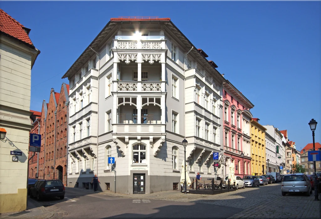 Kamienica o białej elewacji z bogato zdobionymi balkonami, stojąca na skrzyżowaniu ulic w zabytkowej części miasta, otoczona kolorowymi budynkami i zaparkowanymi samochodami, w tle niebieskie niebo.