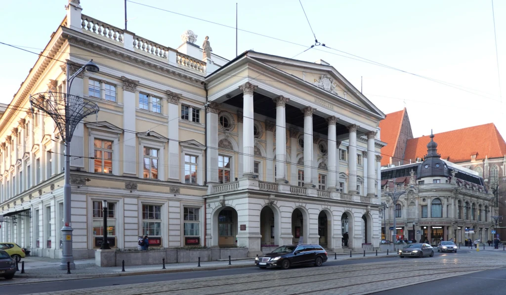 Historyczny gmach z neoklasycystyczną fasadą, ozdobiony wysokimi kolumnami i typowymi dla epoki detalami architektonicznymi. Ulica z tramwajowymi torami oraz pojedynczymi samochodami, widoczna zabudowa sąsiednia z czerwonymi dachami.