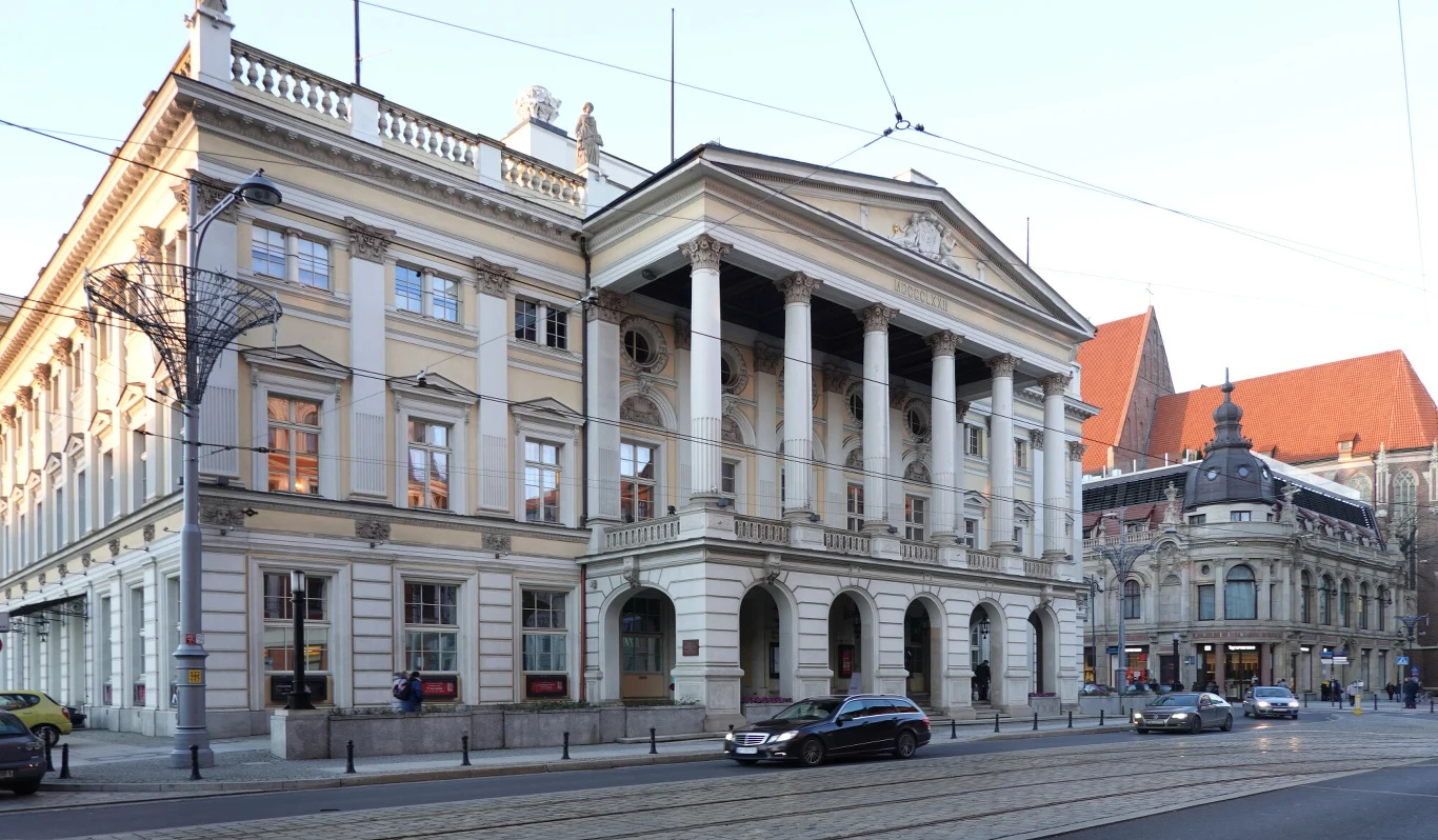 Historyczny gmach z neoklasycystyczną fasadą, ozdobiony wysokimi kolumnami i typowymi dla epoki detalami architektonicznymi. Ulica z tramwajowymi torami oraz pojedynczymi samochodami, widoczna zabudowa sąsiednia z czerwonymi dachami.