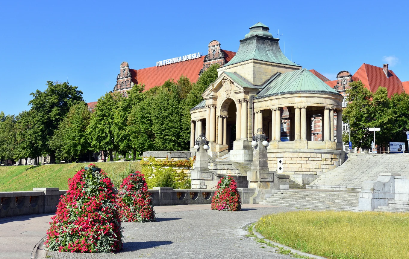 Historyczna rotunda z kolumnami na Wałach Chrobrego w Szczecinie, otoczona kolorowymi kwiatami i zielenią, w tle charakterystyczny gmach Politechniki Morskiej z czerwonym dachem.